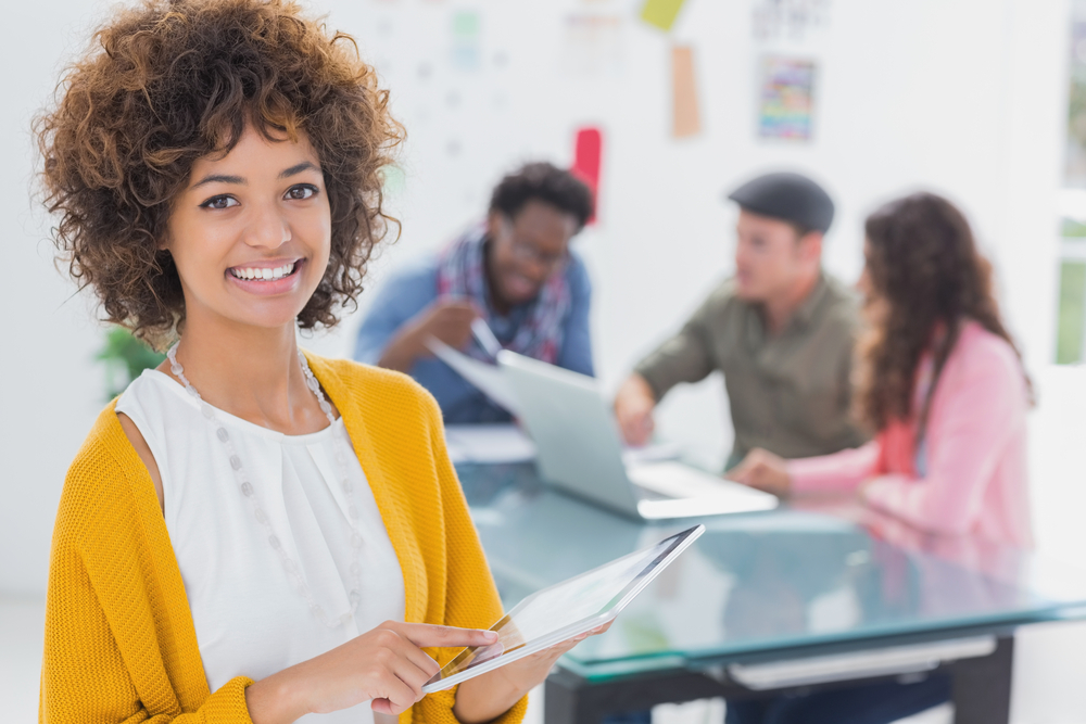 Editor using tablet and smiling at camera as creative team works behind her